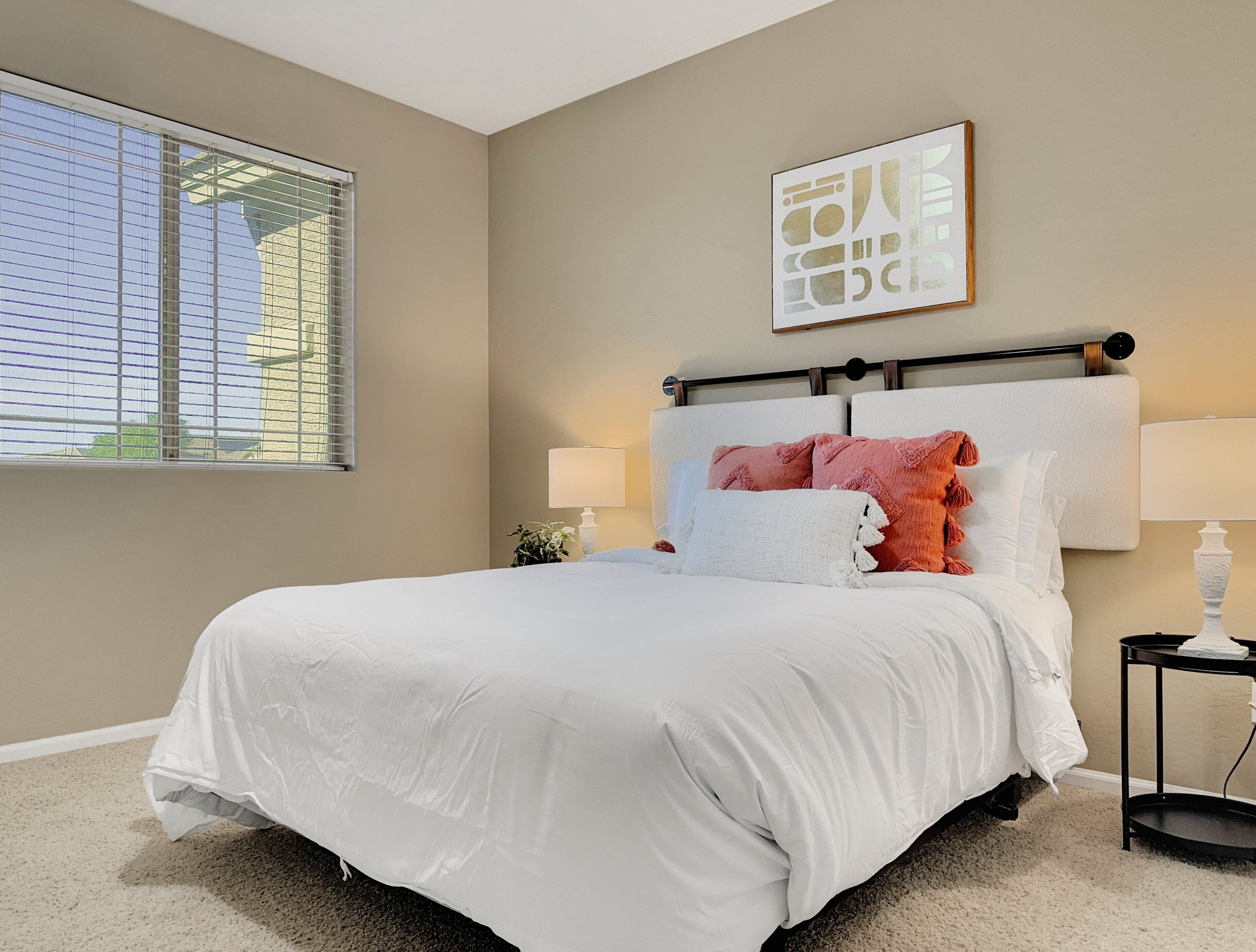 A bedroom with a large bed, two bedside lamps, a window with blinds, and a wall-mounted artwork.