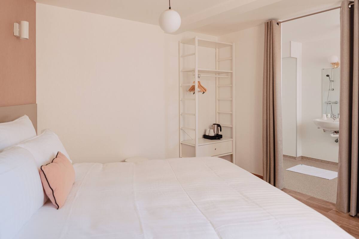 A hotel room with a large bed, a wardrobe, a bathroom with a sink and mirror, and a hanging light fixture.