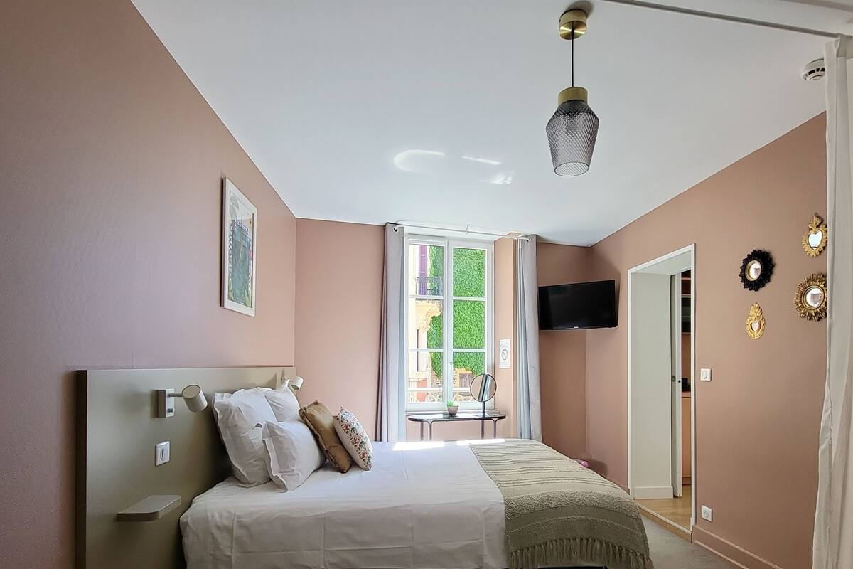 A bedroom with a bed, a bedside table, a window with curtains, a flat-screen TV, and a door leading to another room.