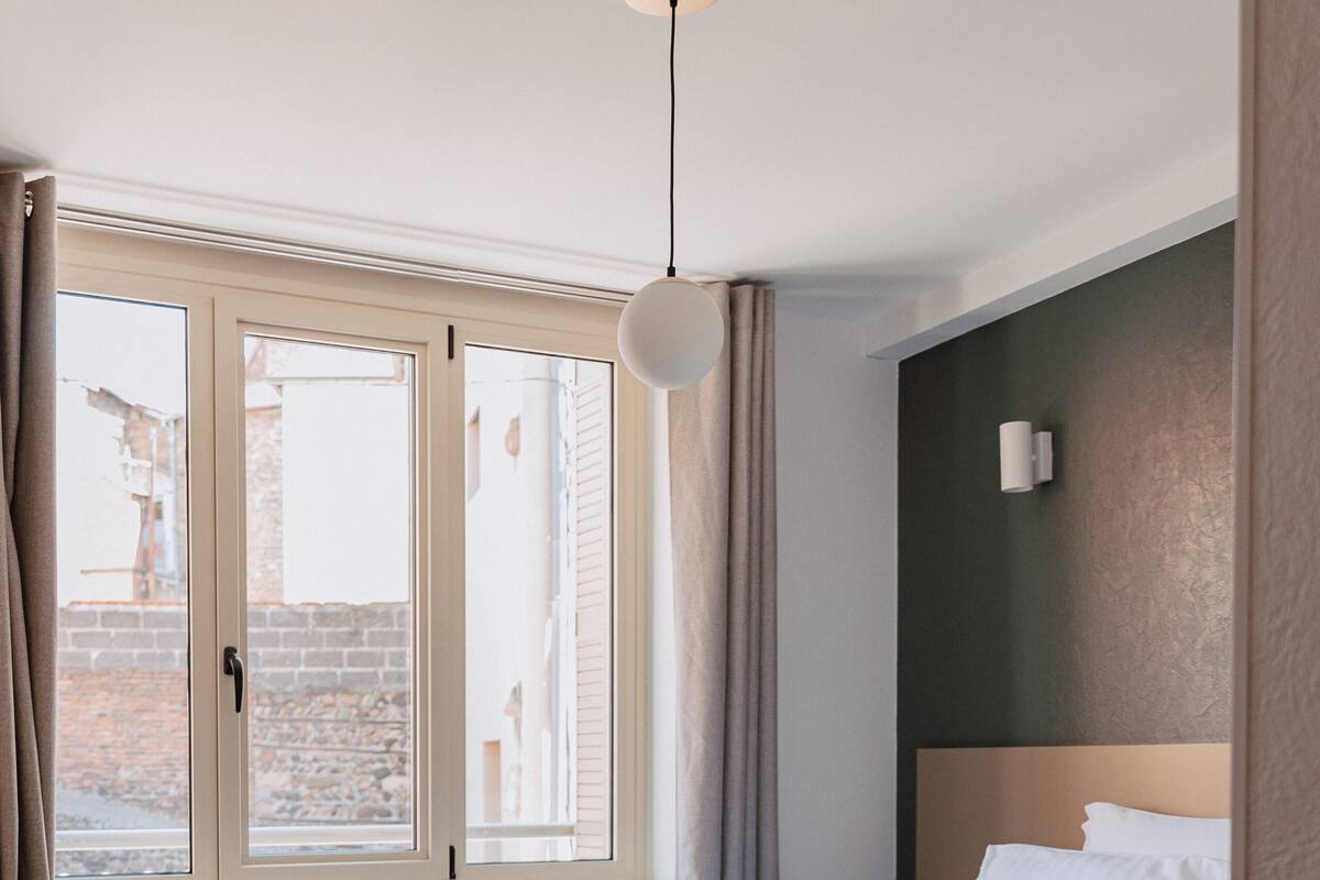 A bedroom with a bed, a window, curtains, a wall-mounted light, and a wooden floor.