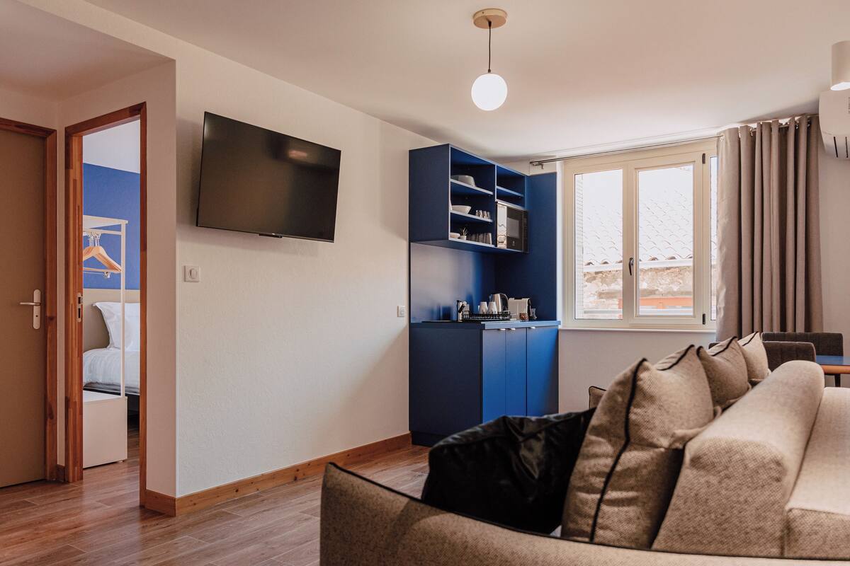 A modern living room with a sofa, a TV mounted on the wall, and a kitchen area with blue cabinets.