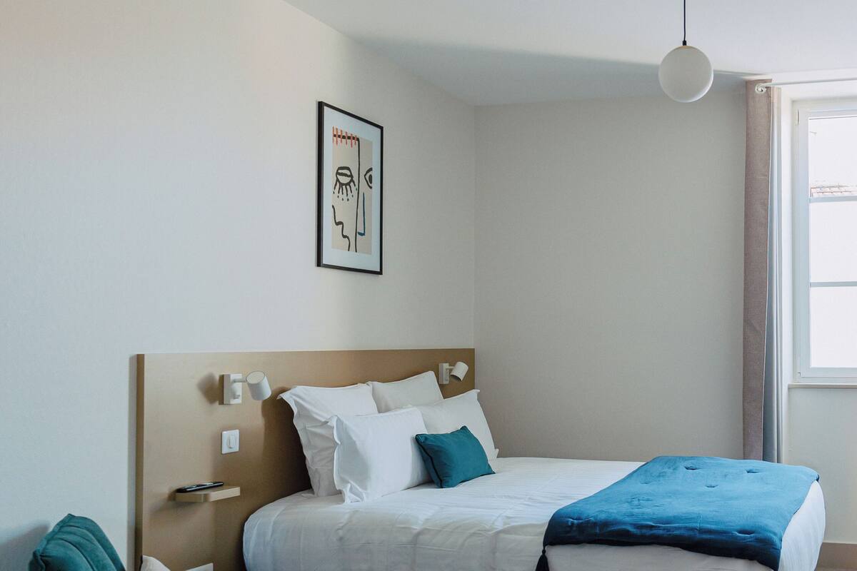 A bedroom with a bed, a chair, a bedside table, a wall-mounted light, and a window.