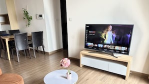 Un saló modern amb televisió de pantalla plana sobre un suport de fusta, zona de menjador amb cadires i una taula rodona petita amb un ram de flors.