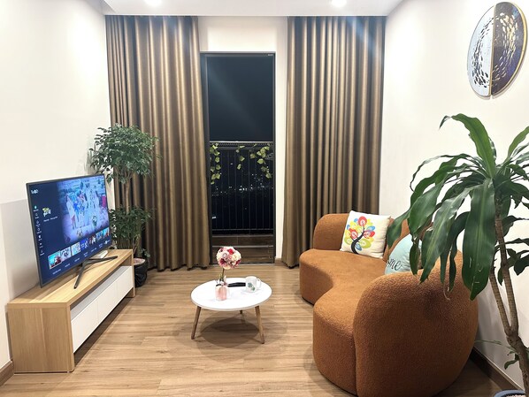 A living room with a flat-screen TV, a round coffee table, a brown sofa, and a large potted plant. - 2 bedrooms apartment 60m2 in Ecopark, nice view, high speed wifi, full amenities (Van Giang District)