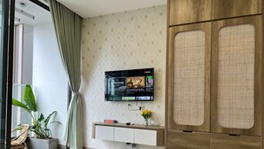 A modern hotel room with a wooden floor, a TV mounted on the wall, a wooden wardrobe, and a potted plant.