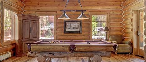 A wooden cabin interior with a pool table, a framed picture on the wall, and a chair by the window.