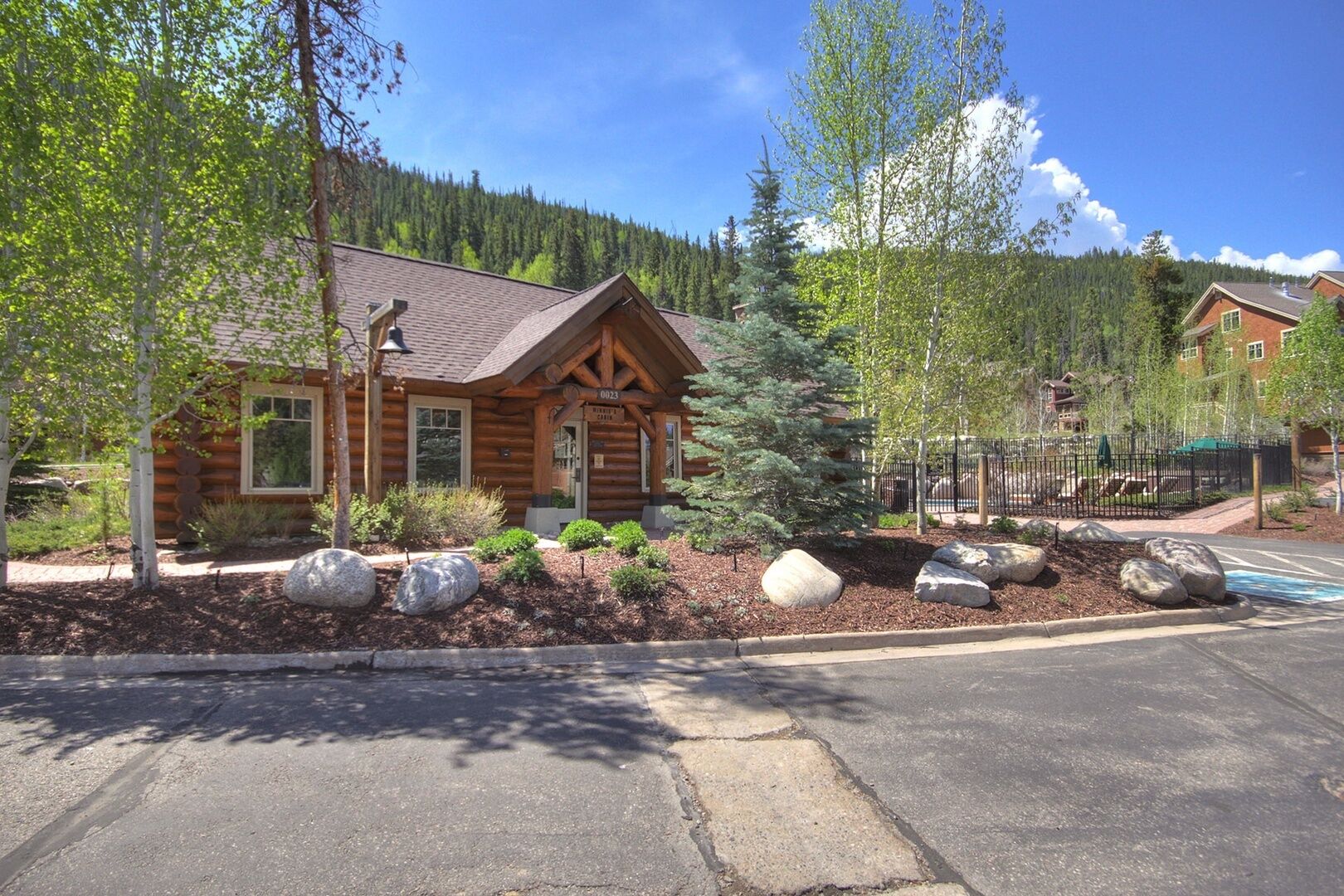 A log cabin with a porch, surrounded by a landscaped garden with rocks and shrubs.