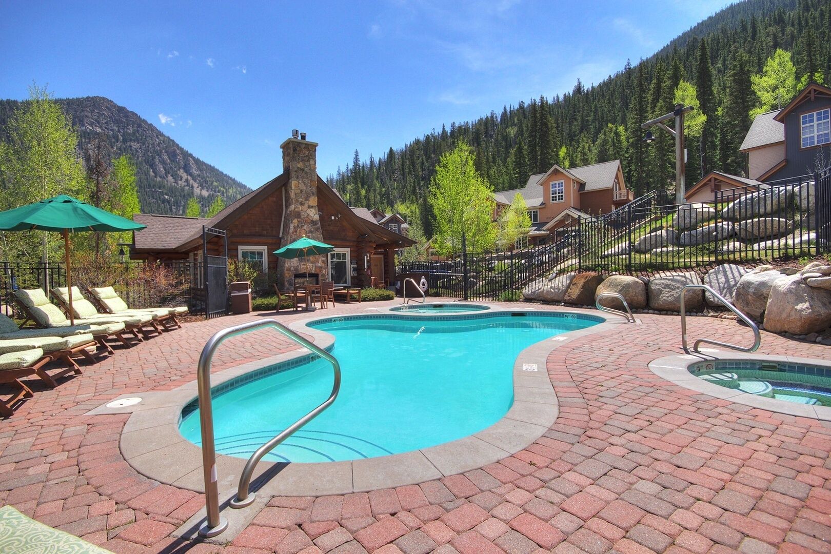A pool area with lounge chairs and umbrellas, surrounded by stone buildings and a mountainous backdrop.
