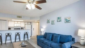 Un salon avec un canapé bleu, une table basse et un ventilateur de plafond. On peut voir au fond quatre tabourets de bar et un coin cuisine.