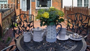 Pöytä on katettu teekupit ja -astiastot sekä kukkakimppu valmiina, ja sitä ympäröi tiiliseinä ja puiset aidat.
