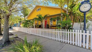 A yellow house with a white picket fence, a clock, and a tree in the foreground. - Charming Ybor City Bungalow with Private Pool Oasis (Tampa)