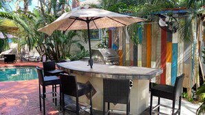 An outdoor bar area with a grill, wicker chairs, and a pool in the background. - Charming Ybor City Bungalow with Private Pool Oasis (Tampa)