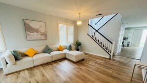 Un salon moderne avec un canapé d’angle, un plancher en bois, un escalier et une cuisine en arrière-plan.