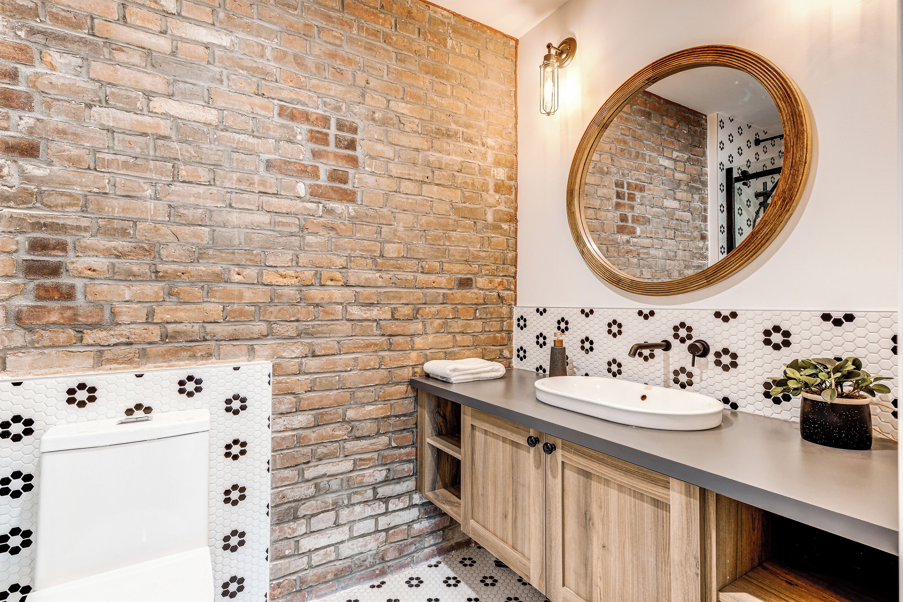 Un baño con pared de ladrillo, un lavamanos blanco, un espejo redondo y una planta en maceta.