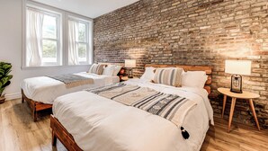 A hotel room with two beds, a brick wall, a large window, and a small table with a lamp. - Bohemian Vieux Quebec Two Bedroom (Québec City)