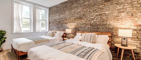 A hotel room with two beds, a brick wall, a large window, and a small table with a lamp.