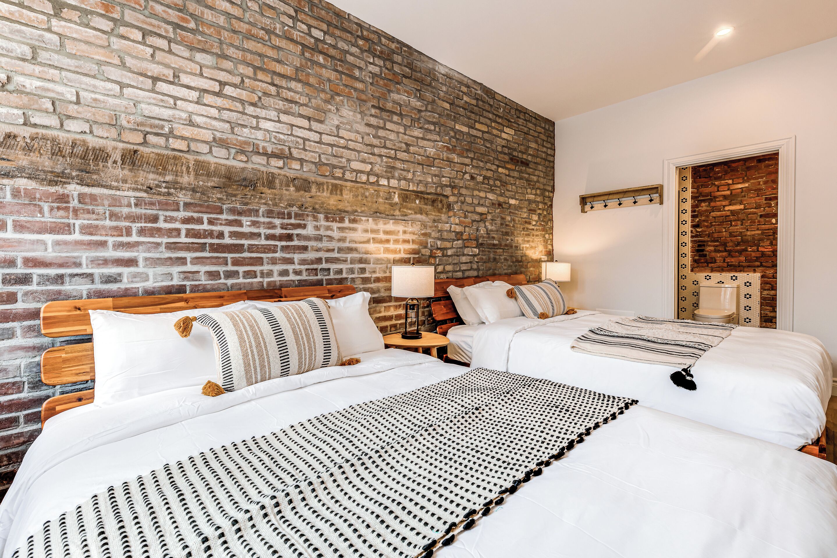 A hotel room with a brick wall, two beds with white bedding, wooden headboards, and bedside tables with lamps.