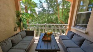 Un balcon avec deux canapés gris, une table en osier sur laquelle est posé un plateau de fleurs, et une vue sur les arbres.