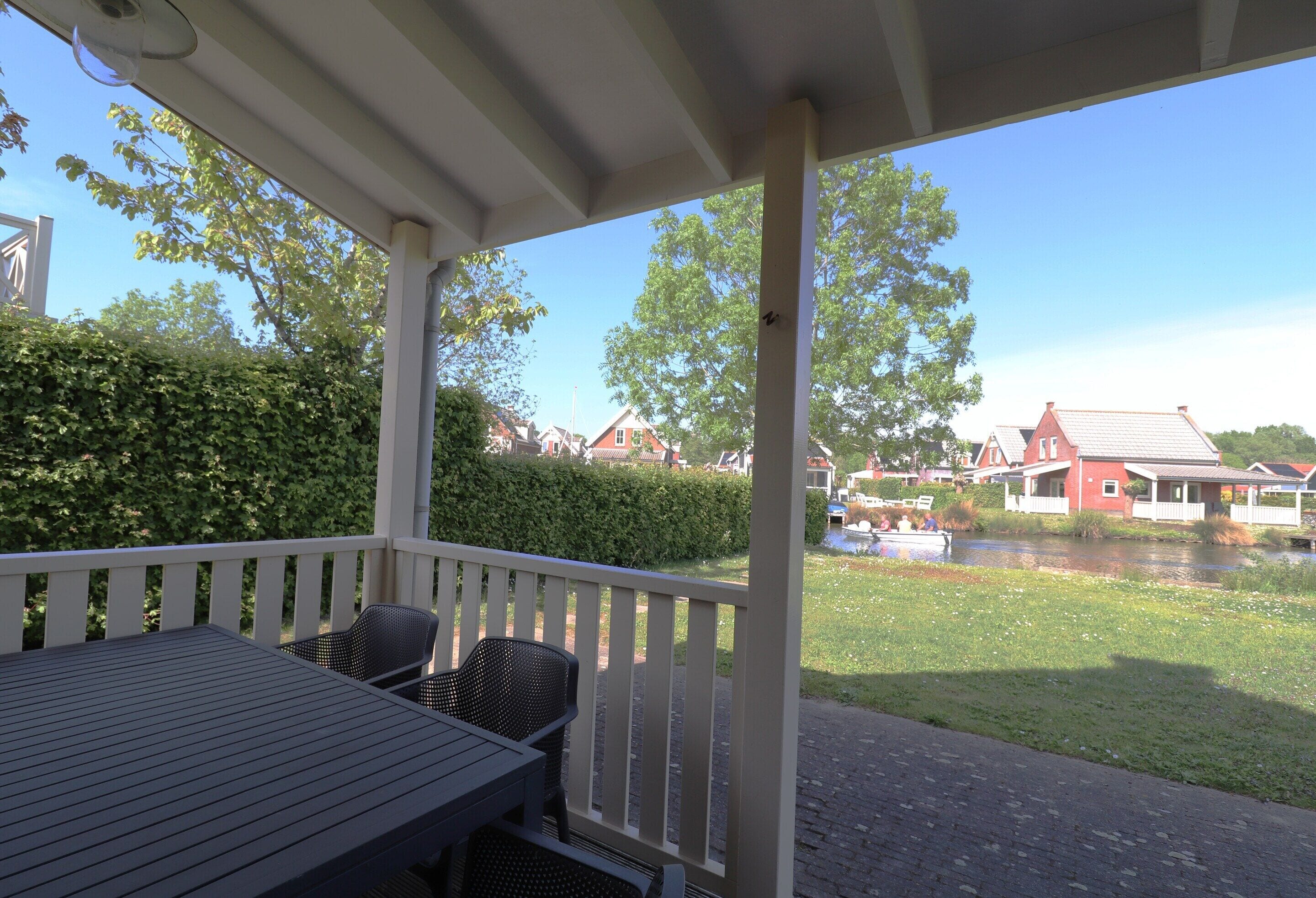 A covered outdoor area with a table and chairs, overlooking a canal with houses and greenery.
