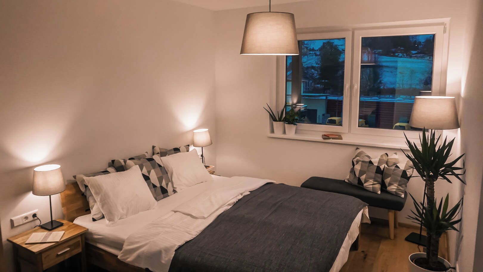 A bedroom with a bed, bedside lamps, a window with plants, and a view of the outside.