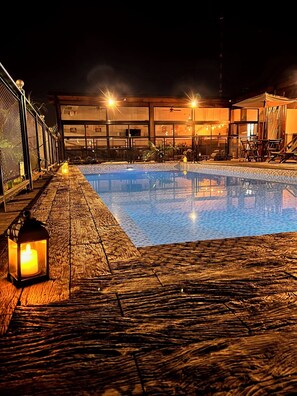 A nighttime swimming pool with a wooden deck, a lantern, and a building in the background. - Alma Pura (Puerto Iguazú)