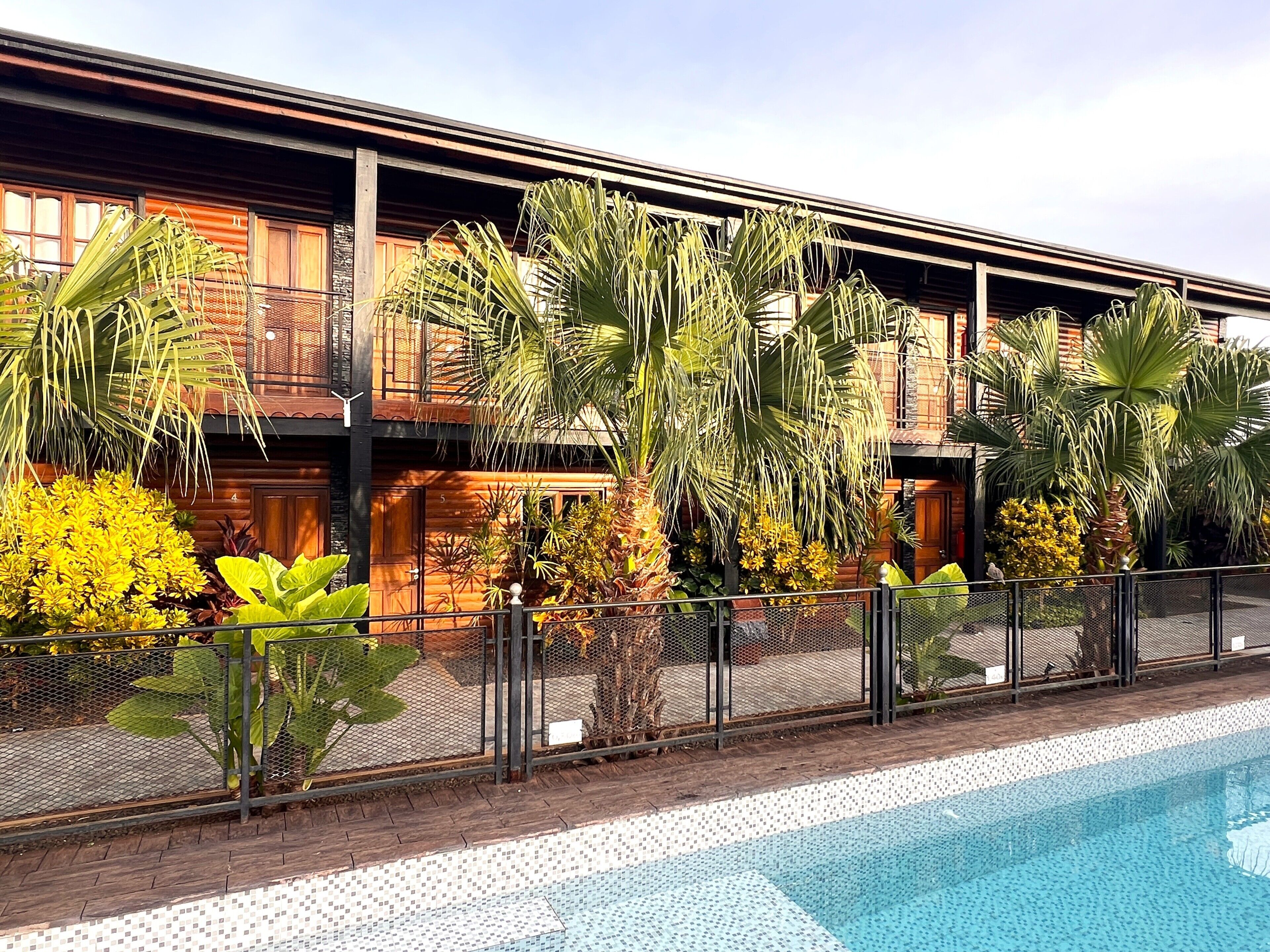 A poolside view of a hotel with a tiled pool, a wooden building with balconies, and various tropical plants.