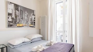 A hotel room with a bed, a window with curtains, a radiator, and a wall-mounted artwork of a cityscape. - Piccole Suite di Charme (Turin)