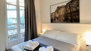 A hotel room with a bed, a window with curtains, a radiator, a wall-mounted painting, and a nightstand with a lamp. - Piccole Suite di Charme (Turin)