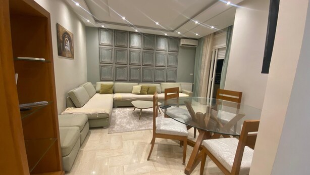 A modern living room with a glass dining table, wooden chairs, a sectional sofa, and a built-in entertainment unit.