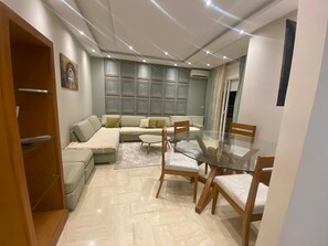 Een moderne woonkamer met een glazen eettafel, houten stoelen, een hoekbank en een inbouwmeubel voor entertainment.