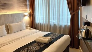 Une chambre d’hôtel avec un lit bien fait, un téléphone sur la table de chevet, une table en verre avec une bouilloire et un plateau contenant divers articles, et une grande fenêtre avec des rideaux.