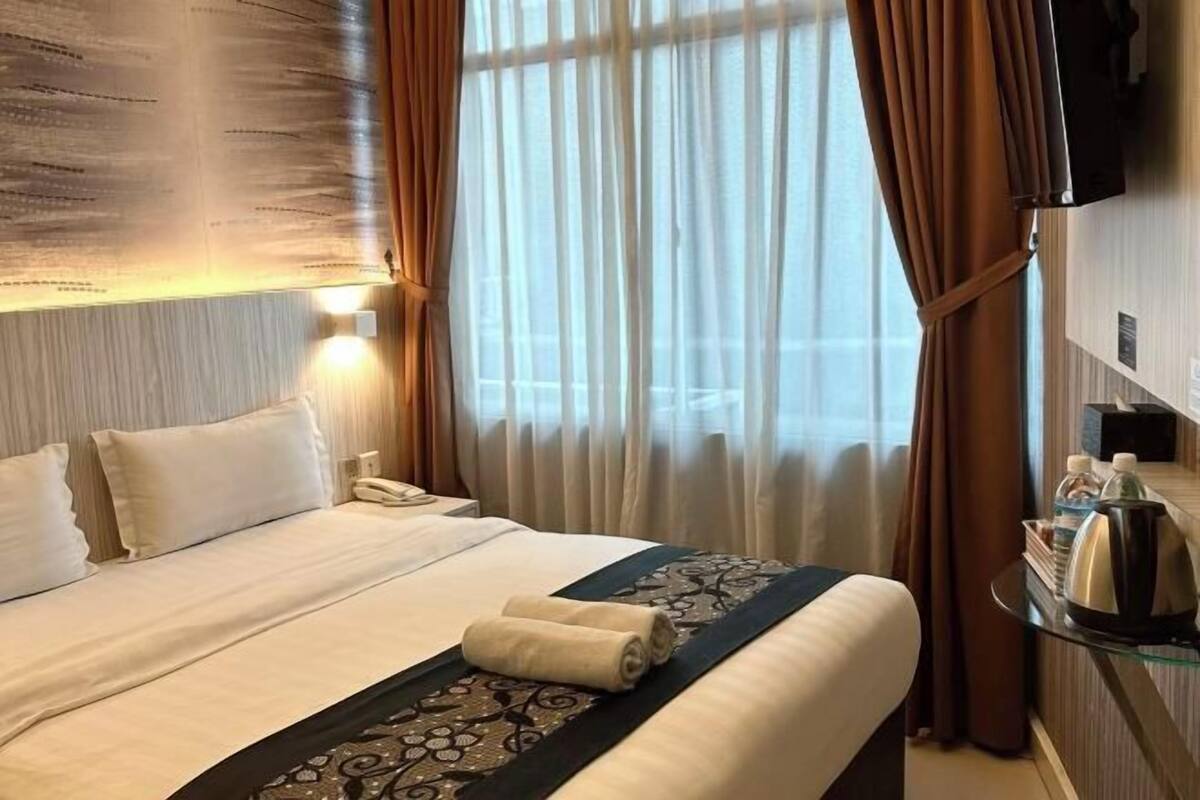 A hotel room with a neatly made bed, a bedside telephone, a glass table with a kettle and a tray of items, and a large window with curtains.