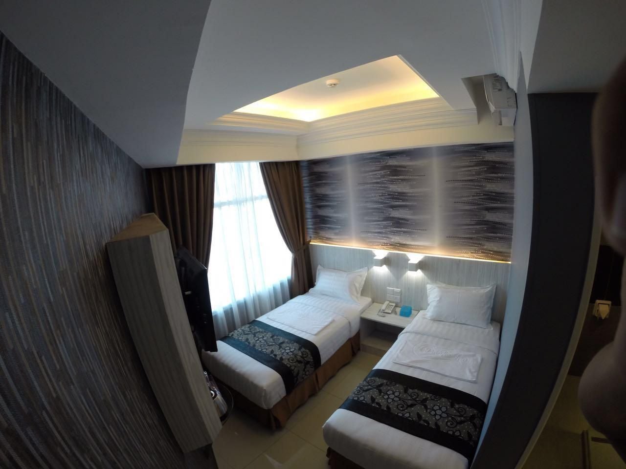 Camera d'albergo con due letti, un'ampia finestra con tende, un condizionatore d'aria a muro e una lampada a soffitto.