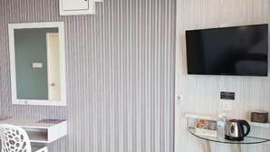 Una habitación con papel tapiz a rayas, un escritorio blanco con una silla, un televisor de pantalla plana montado en la pared y un estante pequeño con un hervidor y otros objetos.