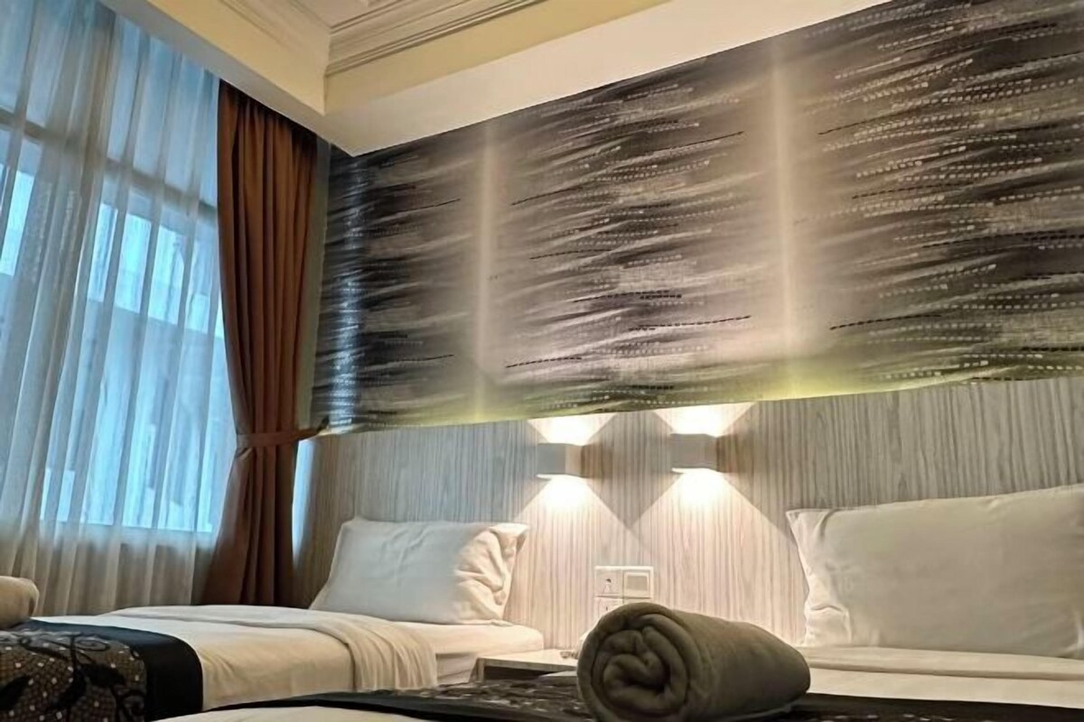 Quarto de hotel com cama, sofá, luminária na parede e janela com cortinas.
