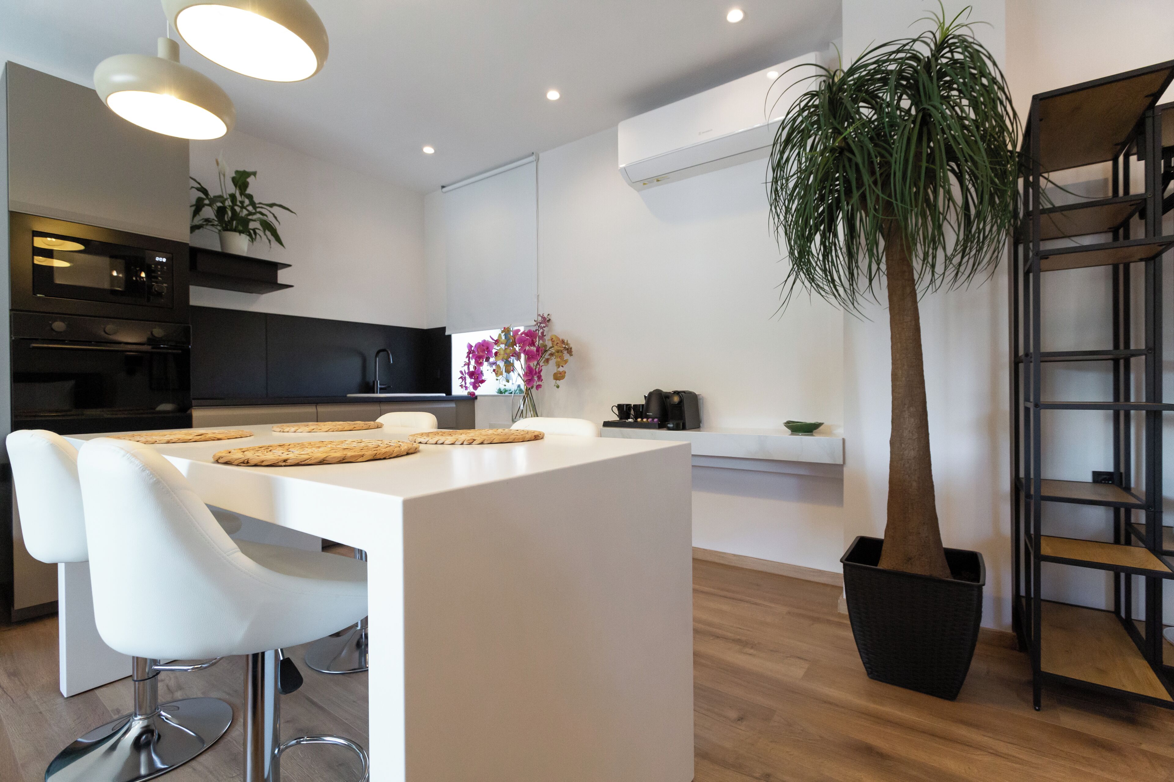 Cozinha moderna com armários brancos, uma ilha central com banquetas, uma planta em vaso e uma estante embutida.