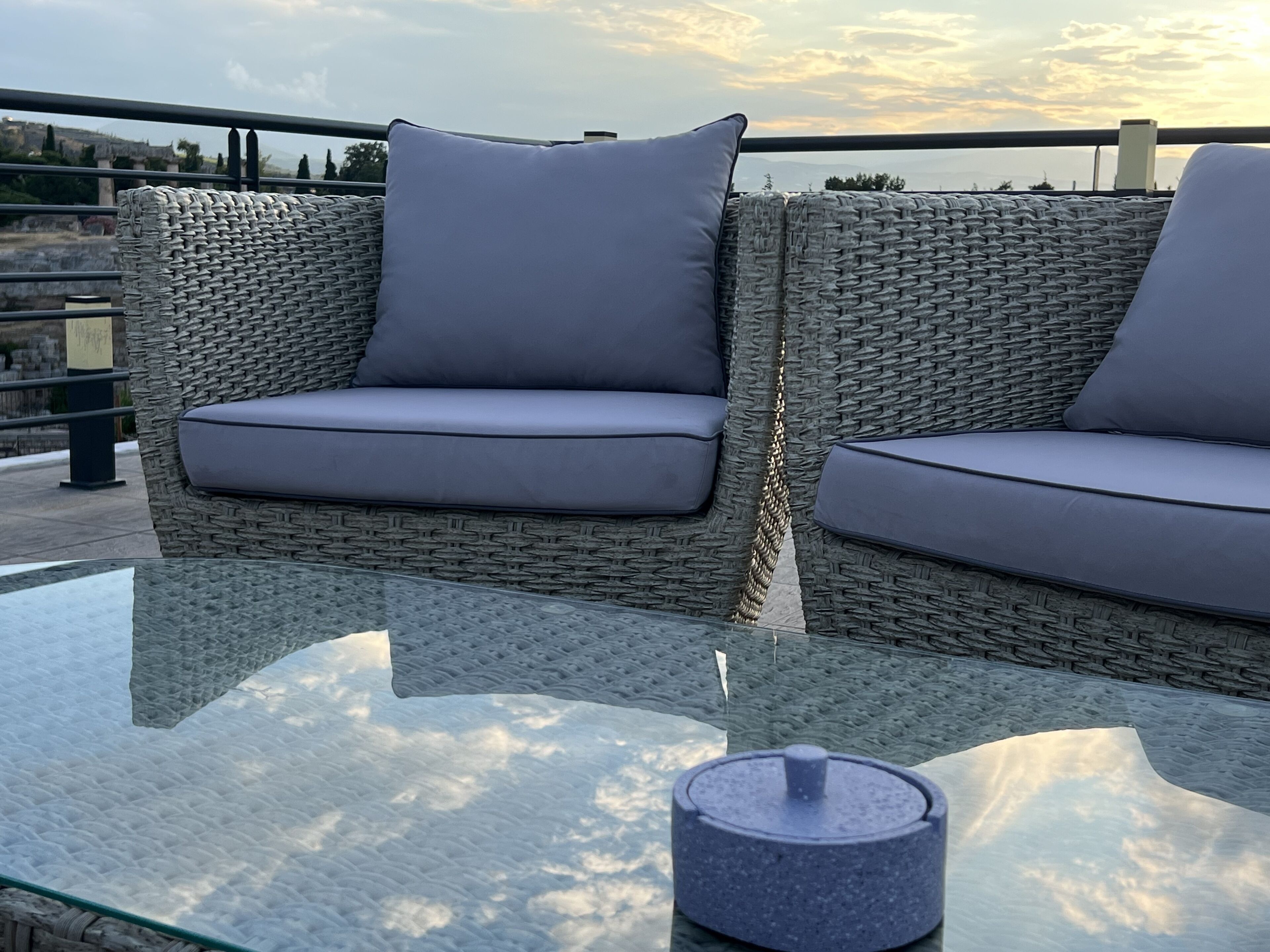 Um sofá de vime com almofadas cinzentas sobre uma mesa de vidro, um objeto azul pequeno sobre a mesa, e um corrimão ao fundo.