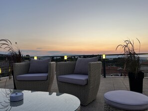 A terrace with wicker furniture and a table, overlooking a cityscape at sunset. - Apollonio Suites (Corinth)