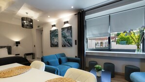 A modern hotel room with a bed, a sofa, a dining table, and a large window with a view of greenery. - Apollonio Suites (Corinth)