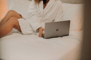 A person in a white robe using a laptop on a bed with white bedding. - Apollonio Suites (Corinth)