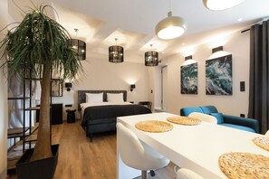A modern hotel room with a bed, a dining table with placemats, a blue sofa, a large potted plant, and artwork on the walls. - Apollonio Suites (Corinth)