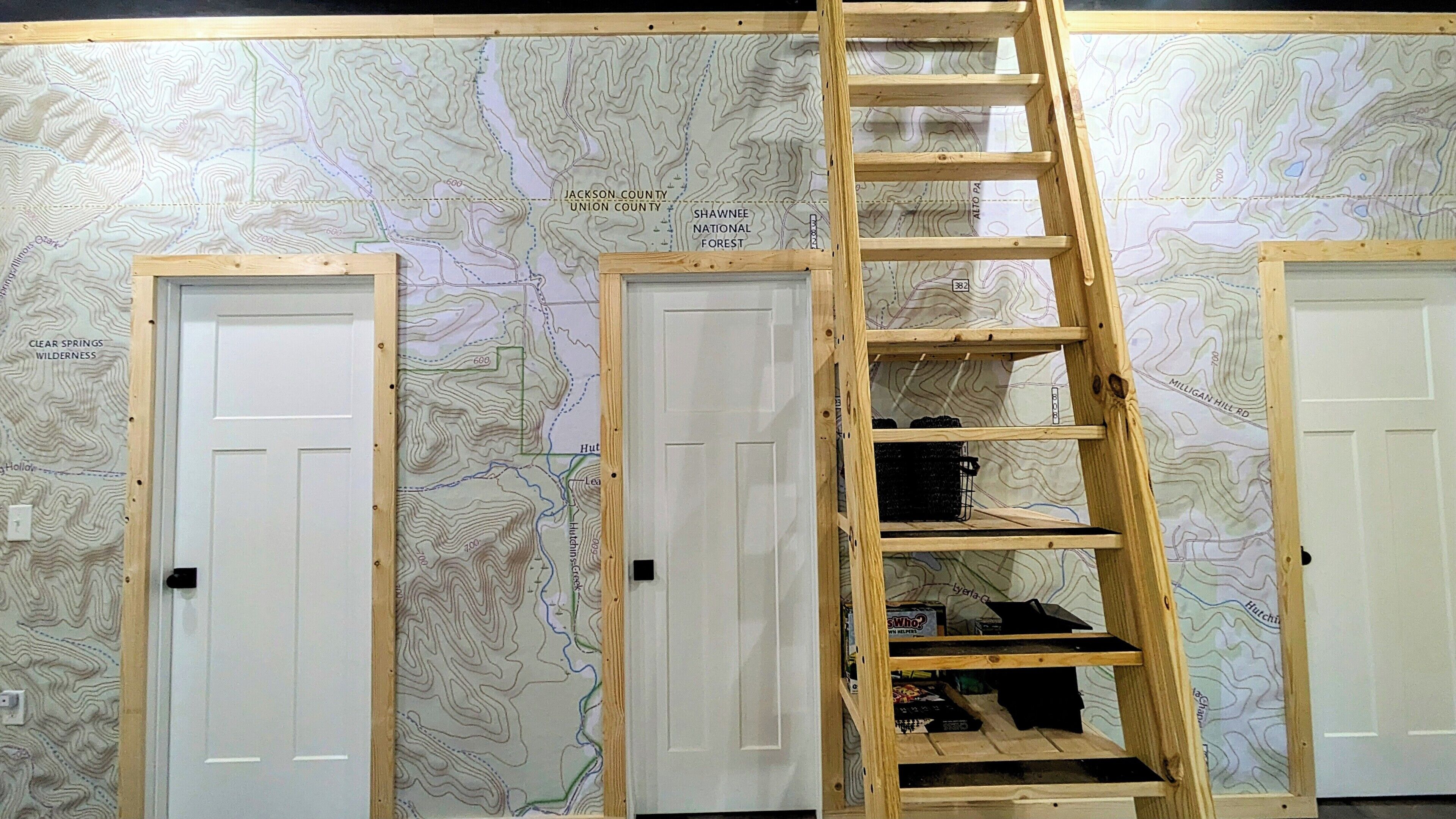 A room with a large topographic map on the wall, a wooden ladder, and three white doors.