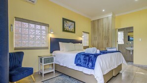 Une chambre à coucher comprenant un lit, des tables de chevet, une chaise, une fenêtre avec des stores, un tableau, et une salle de bain avec un lavabo et un miroir.