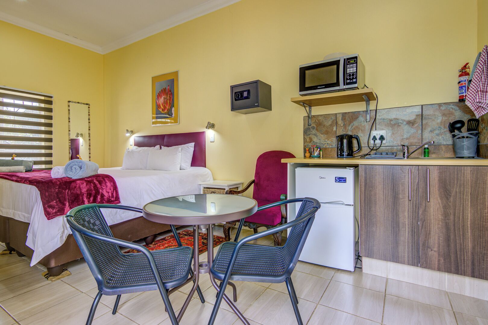 Habitación de hotel con una cama, una pequeña mesa de comedor con sillas, una kitchenette con microondas y un refrigerador, además de una silla roja.