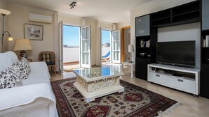 A living room with a sofa, a coffee table, a TV, and a bookshelf. There are two doors leading to a balcony.