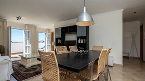 A dining area with a dark wooden table, six wicker chairs, a modern pendant light, and a bookshelf in the background.