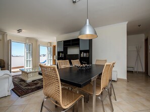Uma sala de jantar com uma mesa de madeira escura, seis cadeiras de vime, uma luminária pendente moderna e uma estante de livros ao fundo.