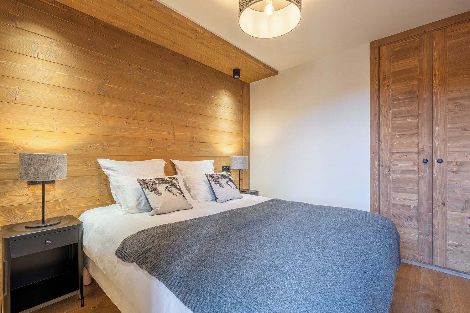 A bedroom with a wooden headboard, a large bed with white bedding, two bedside tables with lamps, and a wooden wardrobe.