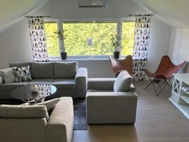 Una sala moderna con un sofá modular, una mesita de centro y una butaca roja. Se observan dos plantas en macetas y una ventana con cortinas.
