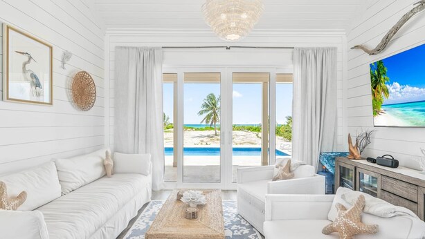 A spacious living room with a large window offering a view of a beach and ocean. There is a white sofa, a coffee table, and a television on a wooden cabinet. - La Dolce Vita Cottage, Beachfront w/ pool directly on Tropic of Cancer Beach! (Moore Hill)
